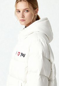 Veste matelassée blanche avec capuche, motif piqué, et texte brodé "I ❤️ YOU" en rouge et noir sur le côté gauche de la poitrine.