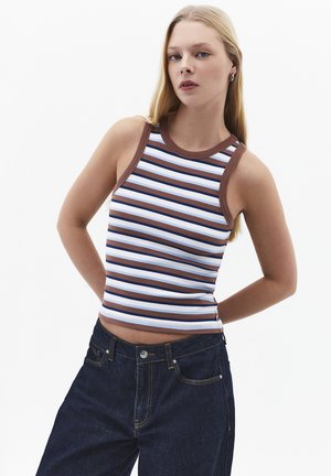 Mujer joven con cabello largo y rubio, vistiendo un top sin mangas a rayas en marrón, blanco y azul, y jeans azul oscuro sobre un fondo blanco.