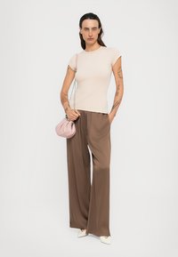 Una donna con tatuaggi indossa una maglietta beige a maniche corte, pantaloni larghi marroni, scarpe bianche e porta una piccola borsa rosa con una catena argentata.