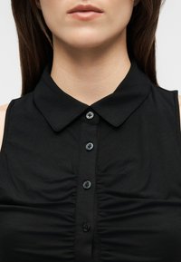 Γυναίκα που φοράει αμάνικο μαύρο πουκάμισο με γιακά, με κουμπιά μπροστά και ρυτιδώσεις στο στήθος.