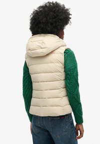 Gilet en couleur crème bouffant avec capuche, présentant des rayures horizontales et une section de manches tricotées vertes. Vue de dos, associé à un jean bleu.