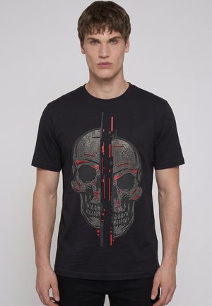 Ung mand iført en sort t-shirt med et pixeleret kranie-grafik opdelt af lodrette røde og grå digitale mønstre, stående mod en ensfarvet baggrund.