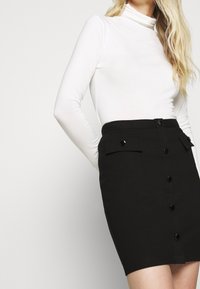 Zwarte kokerrok met knopen aan de voorkant en zijzakken, gemaakt van een gladde stof. Gecombineerd met een aansluitend wit shirt met lange mouwen.