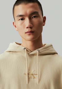 Kreminės spalvos hoodie su raišteliu, turintis subtiliai išsiuvinėtą logotipą krūtinėje. Minkšta medžiaga su lygiu paviršiumi ir laisvesnis fasonas.