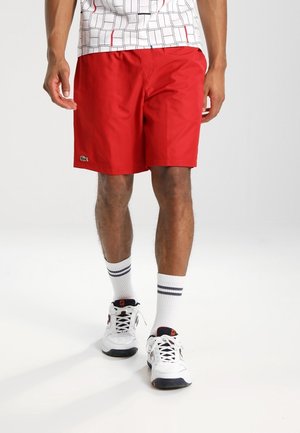 Man draagt rode shorts, witte sokken met zwarte strepen, witte sneakers en een wit overhemd met een zwart-rood ruitpatroon.