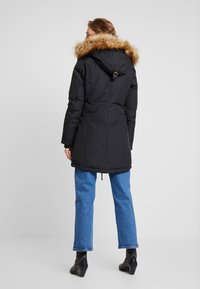 Abrigo parka negro con capucha forrada de piel sintética, que incluye una cintura ajustada y un dobladillo ajustable. Combinado con jeans rectos azules y botas negras de tobillo.