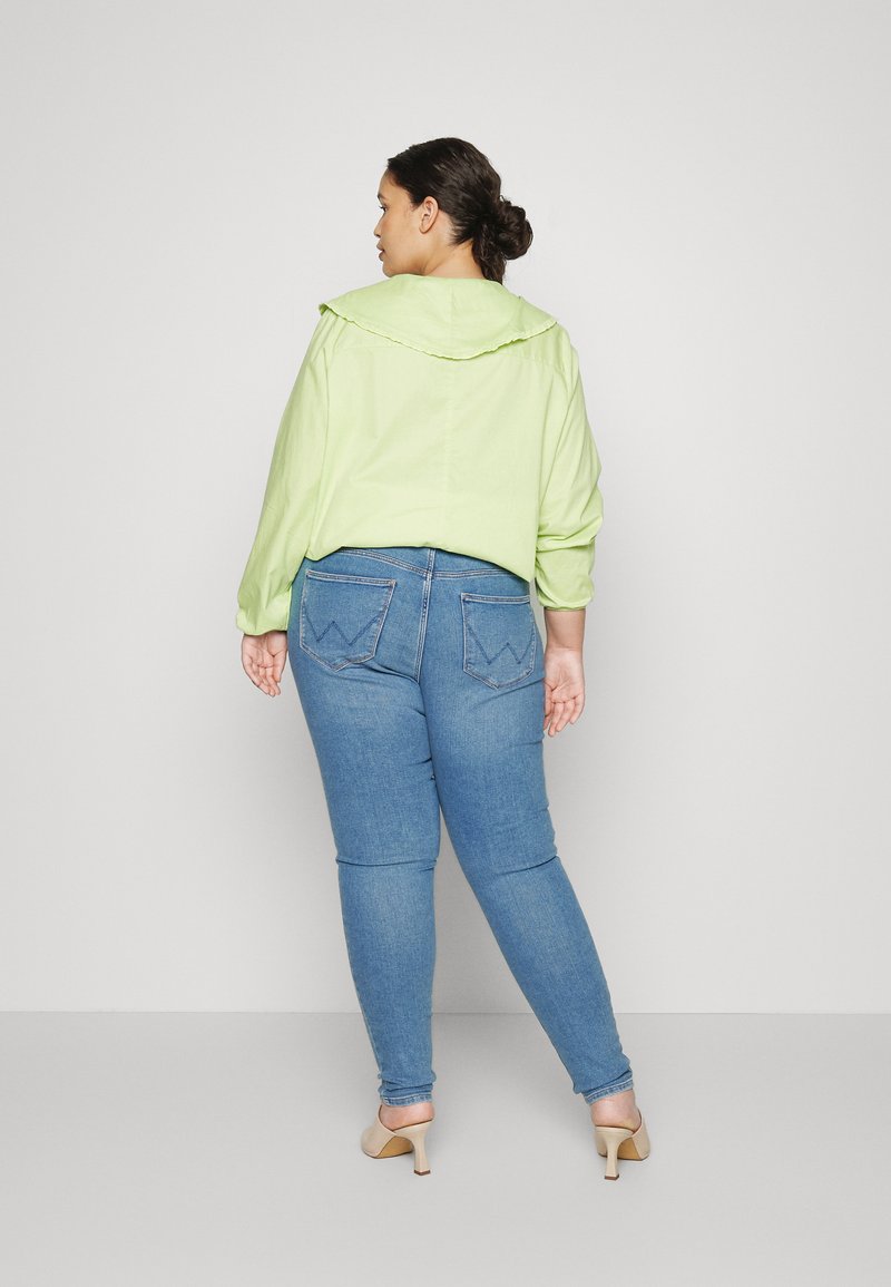 Jasnozielona, lekka koszula z kapturem, w zestawie z dopasowanymi niebieskimi dżinsami z detalami przeszyć tylnych kieszeni, noszona z cielistymi szpilkami.