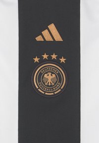 Camiseta deportiva en blanco y negro con un logo de Adidas en dorado bordado y cinco estrellas doradas sobre un emblema circular del Deutscher Fußball-Bund.
