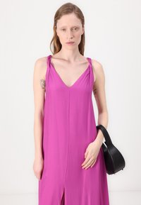 Ροζ αμάνικο φόρεμα με V-λαιμόκοψη και κόμπους στους ιμάντες, που διαθέτει μπροστινό σκίσιμο. Κρατάει μια μικρή μαύρη τσάντα χεριού με καμπυλωτή λαβή.