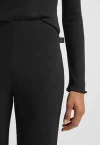 Κοντινό πλάνο ατόμου που φορά μαύρο εφαρμοστό παντελόνι με υφή και αντίστοιχο μακρυμάνικο τοπ, χέρι χαλαρό στο πλάι, μ ε ορατή ετικέτα της μάρκας στο παντελόνι.