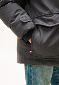 Giacca isolante grigia con una tasca a toppa e un dettaglio logo nero, rosso e bianco sul lato. Visibili jeans in denim blu sottostanti.
