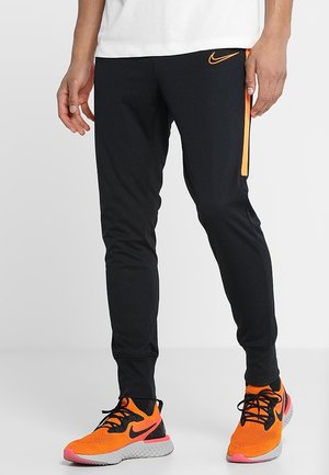 Beine in schwarzen Nike-Trainingshosen mit orangefarbener Seitenstreifen und orangefarbenen Nike-Laufschuhen mit schwarzen Schnürsenkeln und weißen Sohlen.