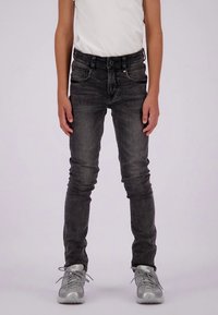 Vaqueros ajustados de color gris oscuro con cinco bolsillos, trabillas para cinturón y una pequeña etiqueta tricolor en la cintura. Combinados con una camisa blanca y zapatillas plateadas.