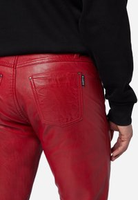 Rode leren broek met een gladde textuur, een aansluitend design en twee achterzakken. Eén zak heeft een klein merklabel. Zwart sweatshirt zichtbaar erboven.