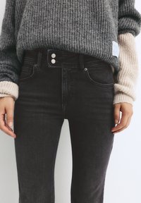 Černé džíny s vysokým pasem a slim fit, které mají dvouknoflíkovou zapínání, pět kapes a mírně vybledlou strukturu. Vyčuhující příjemný šedý pletený svetr.