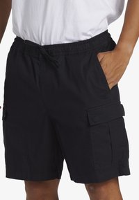 Black cargo shorts with an elastic waistband and adjustable drawstring. Features side pockets and flapped cargo pockets. Lightweight fabric.