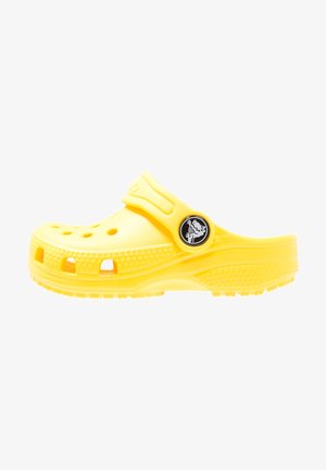 Sabot jaune à enfiler avec des trous de ventilation et une sangle au talon ajustable, arborant un logo circulaire noir avec un design de crocodile blanc.
