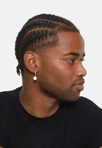 Orecchino a cerchio dorato con pendente in perla bianca. Il soggetto ha i capelli intrecciati e indossa una maglietta nera. Vista di profilo su uno sfondo bianco.