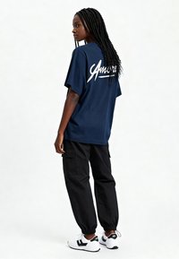 Tumši zilā krāsā pārāk liela T-kreklu ar baltu "Amore" logotipu aizmugurē; apvienots ar melnām biksēm-burām un baltām sporta apavām ar melniem akcentiem.