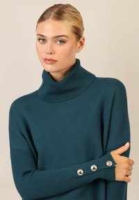 Maglione a dolcevita color turchese con polsini a coste, caratterizzato da dettagli di bottoni decorativi. Tessuto a maglia liscia, design casual e vestibilità rilassata.