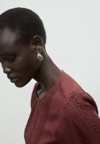 Blusa marrón con detalles de encaje texturizado en los hombros; modelo lleva pendientes de aro dorados; piel suave; fondo neutro.