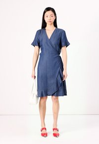 Robe en denim avec des manches courtes et volantées, longueur genou, et volants asymétriques le long de la jupe ; associée à des chaussures rouges et un sac blanc.