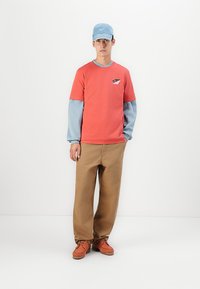 Young man wearing a blue cap, layered red and light blue long-sleeve shirt, tan pants, and brown shoes, standing against a white background.