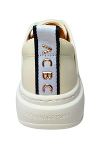 Beige Sneaker mit gepolstertem Fersenbereich, einem weiß-schwarz gestreiften Etikett und metallischen Akzenten. Die Sohle ist dick und strukturiert und trägt den Markennamen.