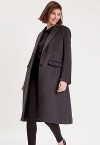 Grahamfarbener Wollmantel mit maßgeschneiderter Passform, einreihiger Vorderseite, zwei Knöpfen und seitlichen Taschen. Der Stoff wirkt weich und strukturiert.