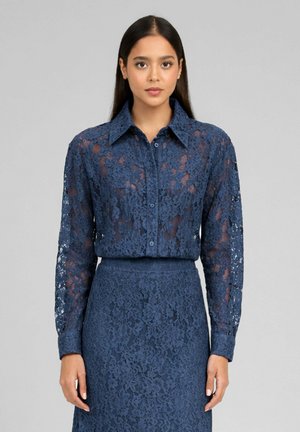 Femme portant une chemise blanche à manches longues en dentelle bleu marine rentrée dans une jupe taille haute assortie, debout devant un fond gris uni.