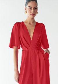 Kvindeligt med mørkt hår i en slicked-back frisure bærer en rød jumpsuit med dyb V-udskæring, pufærmer til albuen og guld hoop-øreringe.