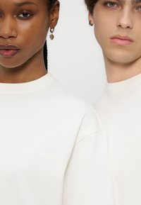 Κοντινό πλάνο δύο ατόμων που φορούν λευκές φούτερ, το ένα με σκουλαρίκια σε σχήμα καρδιάς και το άλλο με σκουλαρίκια σε σχήμα αστεριού.