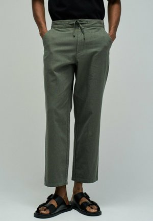 Homme portant un pantalon à cordon olive et des sandales noires à boucle, les mains dans les poches, debout devant un fond gris uni.