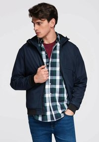 Námořnická kapucová bunda přes zelenou a bílou kostkovanou košili, s tmavými manžetami a zipem vpředu. Model stojí čelom слегка na stranu.