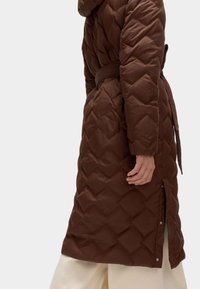 Brun quiltet lang frakke med hætte, bælte i taljen, sideslids og trykknaplukninger, lavet af glat, slidstærkt materiale.