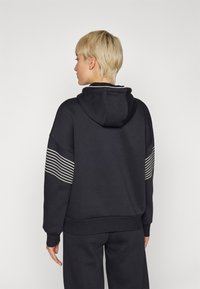 Svart hettegenser med kengurulomme, med hvite horisontale striper på ermene. Laget av en bomullsblanding, har en løs passform.