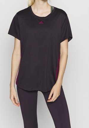 Vrouw draagt een donker kortärmelig Adidas T-shirt met roze logo en bijpassende donkere leggings, staand tegen een effen achtergrond.
