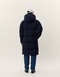 Uomo che indossa un lungo cappotto in nylon blu scuro con cappuccio, jeans blu, scarpe nere e un cappellino da baseball grigio chiaro, in piedi di spalle.