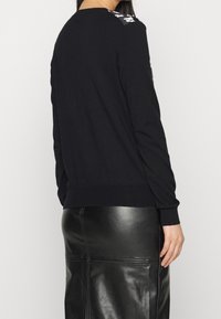 Žena v černém svetru s dlouhým rukávem a bílými vzory na rameni a v černé kožené sukni, otočená zády ke kameře.