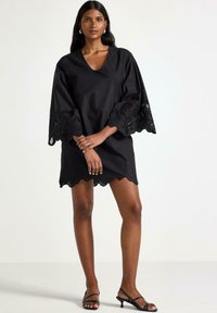 Svart klänning med V-ringning och vida ärmar som har intrikata spetsdetaljer. Kjolens nederkant har en scallopkant. Bäraren står i sandaler.