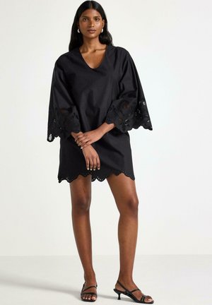 Svart klänning med V-ringning och vida ärmar som har intrikata spetsdetaljer. Kjolens nederkant har en scallopkant. Bäraren står i sandaler.