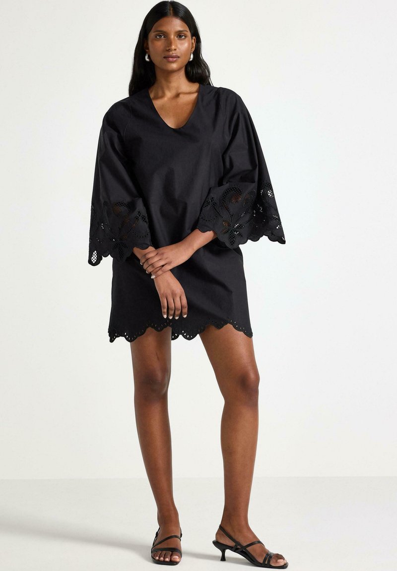 Svart klänning med V-ringning och vida ärmar som har intrikata spetsdetaljer. Kjolens nederkant har en scallopkant. Bäraren står i sandaler.