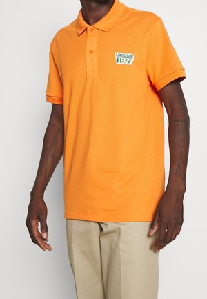 Mann trägt ein orangefarbenes Poloshirt mit kurzen Ärmeln und dem "Lacoste 1927"-Logo auf der Brust sowie beige Hosen. Seine Unterarme sind tätowiert.
