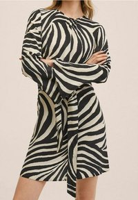 Femme les bras croisés portant une robe noire et blanche à rayures zébrées avec des manches longues évasées et une ceinture nouée à la taille.