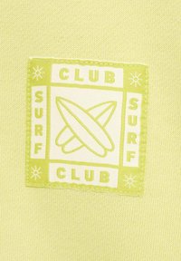 Tessuto verde chiaro con una macchia bianca quadrata che presenta la scritta "SURF CLUB" e delle tavole da surf incrociate. La cucitura è visibile lungo i bordi.