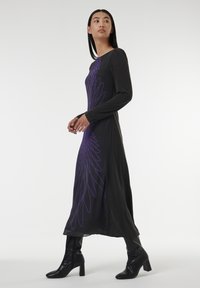 Robe noire à manches longues avec un motif graphique violet ressemblant à des plumes, fabriquée dans un tissu lisse, associée à des bottes à talons noires montantes.