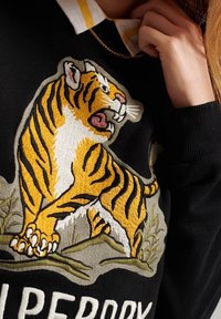 Svart sweatshirt med en broderad tiger i gult och vitt med en grön och beige bakgrund, som är ihop med en skjorta med krage under.