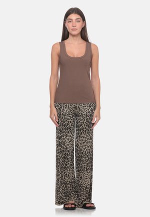 Donna in piedi che indossa una canotta marrone senza maniche, pantaloni a gamba larga con stampa leopardata e sandali neri su sfondo bianco semplice.