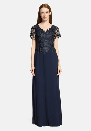 Femme portant une robe longue bleu marine avec des manches courtes en dentelle florale transparente et un corsage supérieur, debout devant un fond clair uni.