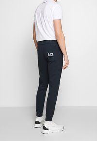 Tmavě modré teplákové kalhoty s elastickým pasem a manžetami, s zadní kapsou s bílým logem EA7. Kombinované s bílým tričkem s kulatým výstřihem.